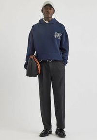 Felpa navy con logo frontale, pantaloni neri e scarpe nere. Il modello porta una borsa nera con accenti marroni. Un cappello grigio completa il look.