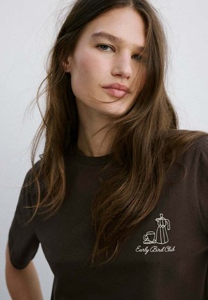 Jeune femme aux longs cheveux bruns porte un t-shirt foncé avec une broderie blanche d'une cafetière et d'une tasse "Early Bird Club" sur la poitrine.