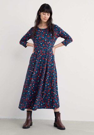 Kvetinové midi šaty v modrej farbe s červenými a bielymi vzormi, okrúhlym výstrihom, rukávmi po tri štvrtiny a rozšírenou sukňou. Hnedé členkové čižmy dotvárajú vzhľad.