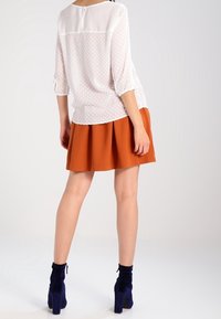 Blouse blanche transparente avec des pois texturés, associée à une jupe plissée orange. Des bottines bleues à la cheville complètent la tenue.