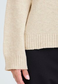Détail de manches d'un pull en tricot beige avec un poignet côtelé, montrant une texture douce. L'arrière-plan présente un vêtement noir.