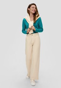 Helle beige Kick-Flare-Hosen mit zwei Knöpfen, kombiniert mit einem türkisfarbenen Langarmshirt und einem cremefarbenen Pullover, der lässig um die Schultern gebunden ist.