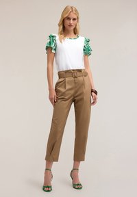 Top bianco con maniche arricciate floreali verdi, abbinato a pantaloni beige a vita alta con cintura e sandali verdi con cinturini. Texture morbide.