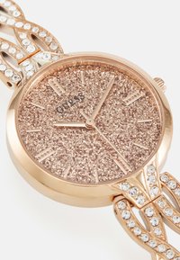 Orologio rosa oro con un quadrante rosa oro scintillante, cinturino a maglie con cristalli trasparenti e cassa rotonda. Inclusi marker orari numerici.