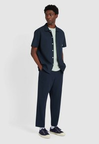 Shirt navy a maniche corte con colletto button-down, t-shirt verde menta sotto, pantaloni testurizzati blu scuro e sneakers nere con suole crema.