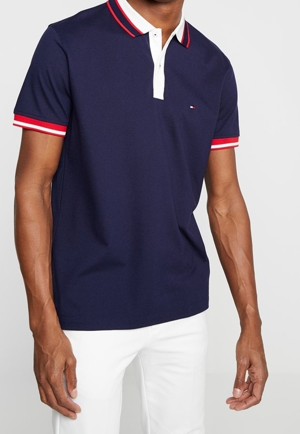 Homme portant un polo bleu marine avec un col et des manches rayés rouge et blanc, associé à un pantalon blanc, debout devant un fond uni.