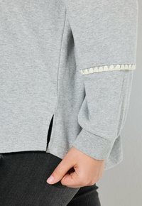 Šedý dlouhý rukáv s texturovaným vzorem na lemu rukávu a bočními rozparky, vyrobený z měkké látky. Kombinace s tmavými džíny.