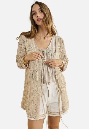 Joven mujer atando los cordones de un cárdigan beige de punto abierto, llevando una camiseta sin mangas a rayas y pantalones cortos blancos con flecos sobre fondo blanco.