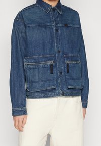 Jeansjacka i mörkblått med knäppning fram, två framfickor med dragkedja och skjortkrage. Har en strukturerad yta och kontrasterande sömmar.