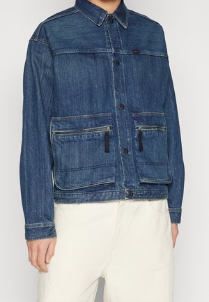 Giacca di denim di colore blu scuro con chiusura a bottoni, due tasche frontali con zip e colletto da camicia. Presenta una finitura testurizzata e cuciture a contrasto.