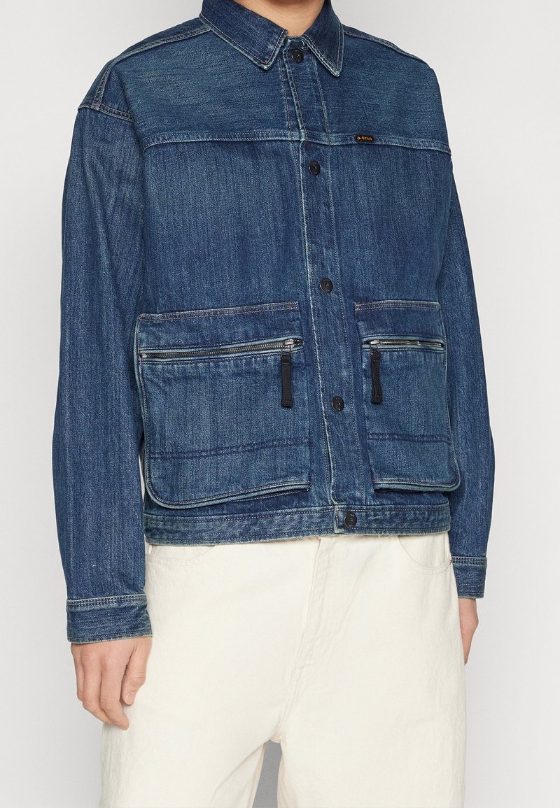Jeansjacka i mörkblått med knäppning fram, två framfickor med dragkedja och skjortkrage. Har en strukturerad yta och kontrasterande sömmar.