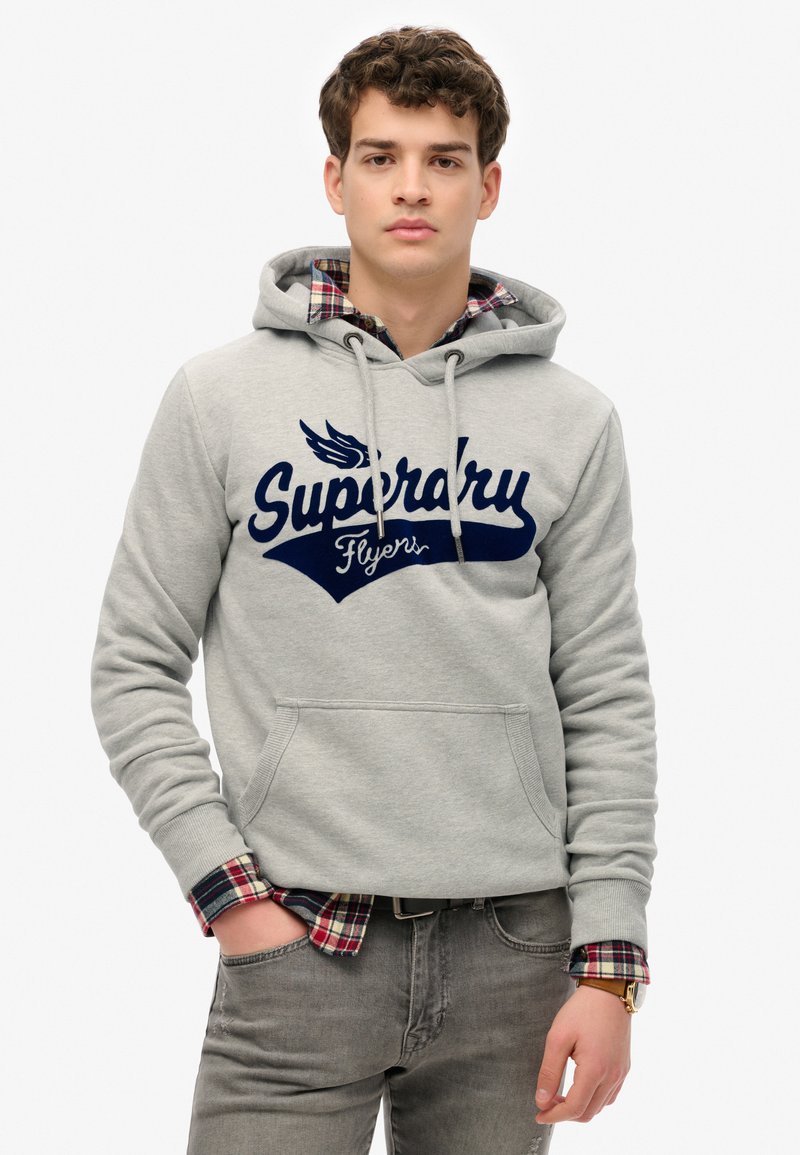 Felpa grigia con logo navy "Superdry Flyers", tasca a marsupio frontale e cappuccio con cordino. Sovrapposta a una camicia a quadri rossa nei polsini.