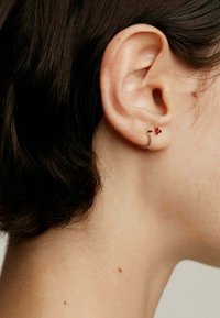 Pendientes de metal en tono dorado con diseño de serpiente y un pequeño acento de gema roja, colocados en una oreja perforada. Primer plano de la oreja y peinado.