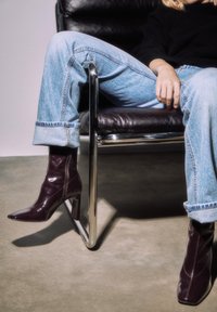 Stivaletti alla caviglia in pelle verniciata color bordeaux con punta affilata e tacco largo, indossati con jeans celesti arrotolati, seduti su una sedia nera.