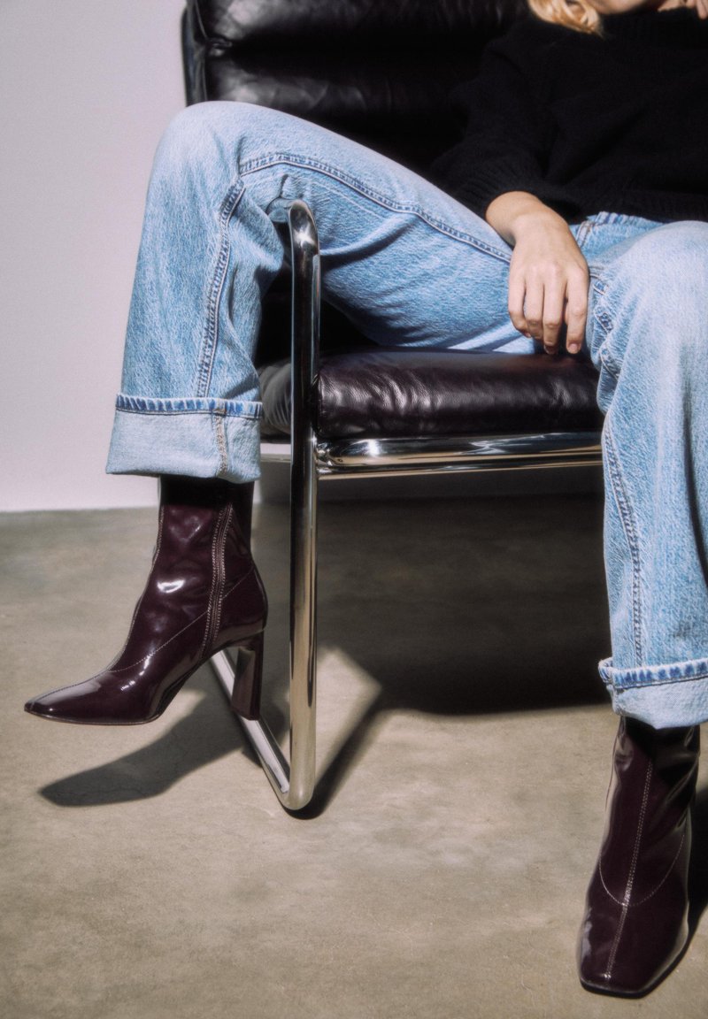 Stivaletti alla caviglia in pelle verniciata color bordeaux con punta affilata e tacco largo, indossati con jeans celesti arrotolati, seduti su una sedia nera.
