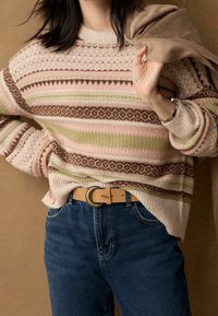 Pull en maille beige avec des rayures roses, vertes et marron ; motifs texturés ; associé à un jean bleu et une ceinture marron clair.