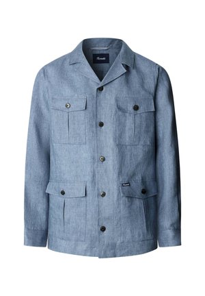 Mann trägt eine hellblaue Jacke mit Knöpfen und vier Klappentaschen über einem weißen Hemd, steht vor einem einfarbigen hellen Hintergrund.