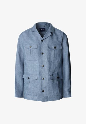 Hombre con chaqueta azul claro abotonada con cuatro bolsillos de solapa sobre una camisa blanca, de pie contra un fondo liso y claro.