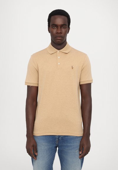 Beige pikétrøye med krage, tre knapper og en liten brodert logo på venstre bryst. Korte ermer, myk bomullsblanding.