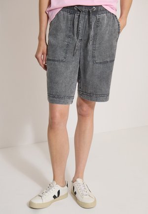 Personne portant un short en jean noir délavé jusqu'aux genoux avec de grandes poches avant et des baskets blanches avec des détails noirs.