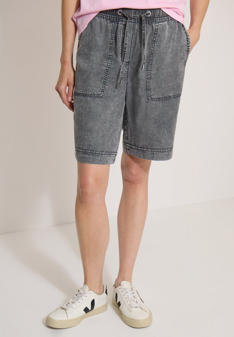 Personne portant un short en jean noir délavé jusqu'aux genoux avec de grandes poches avant et des baskets blanches avec des détails noirs.