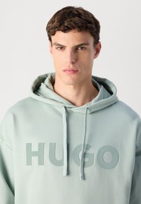 Giovane uomo con capelli corti castani che indossa una felpa leggera verde con la scritta "HUGO" stampata davanti, che guarda dritto davanti a sé con un'espressione neutra.