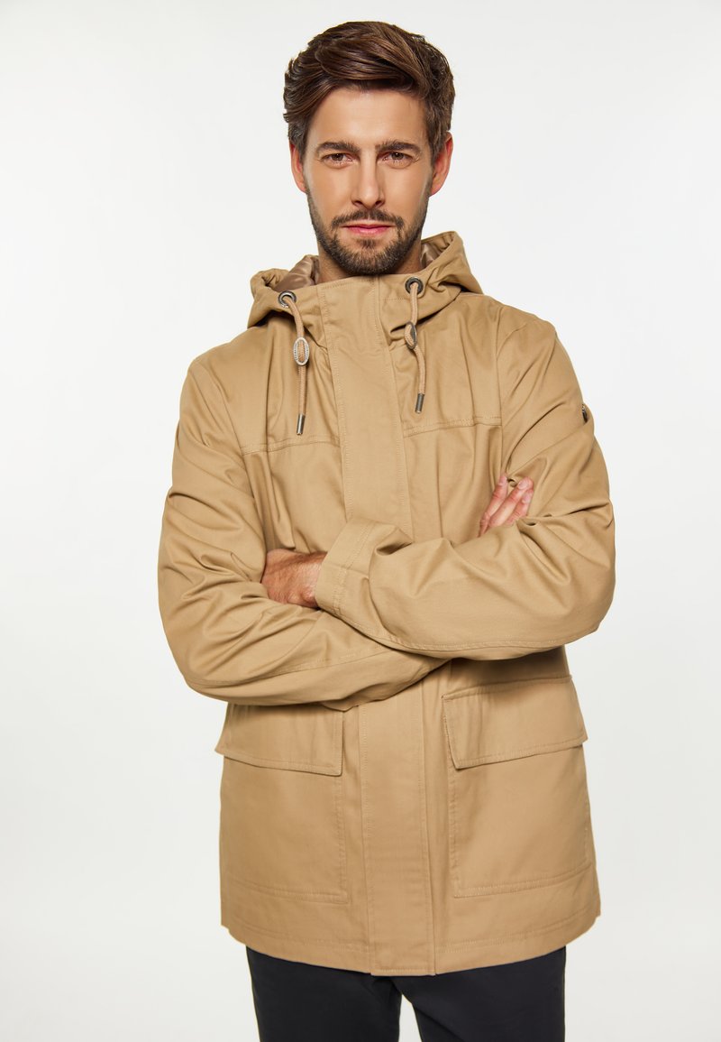Tan huva jacka med dragkedja fram, två fickor och en texturerad yta. Designen har justerbara snörstängningar på huvan.
