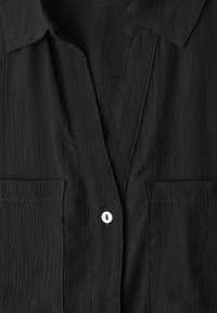 Chemise noire texturée avec col, deux poches poitrine et un seul bouton blanc fermé près du centre.