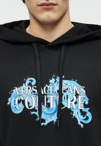 Κοντινό πλάνο ενός ατόμου που φοράει μαύρο φούτερ με κουκούλα με το κείμενο "Versace Jeans Couture" και μπλε διακοσμητικά στροφικά μοτίβα στο στήθος.