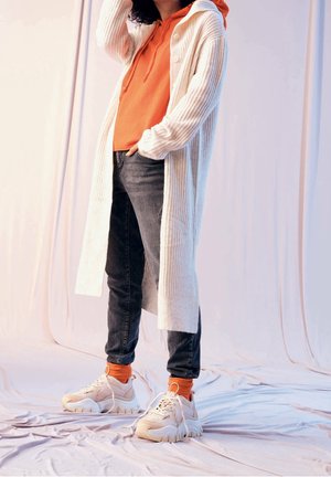 Pitkä, poimutettu valkoinen neuletakki oranssin hupullisen paidan päällä, yhdistetty tummiin farkkuihin ja paksuihin valkoisiin tennareihin, joissa on oranssit sukat korostuksena.