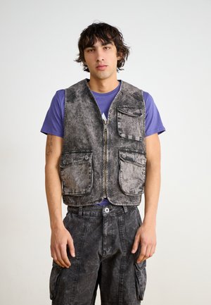 Gilet in denim grigio scolorito con zip frontale, quattro tasche anteriori e design senza maniche, indossato sopra una t-shirt viola e pantaloni corti neri.