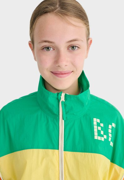 Jeune personne aux yeux et cheveux clairs, portant une veste à fermeture éclair verte et jaune avec un texte blanc partiel sur la poitrine, avec un léger sourire