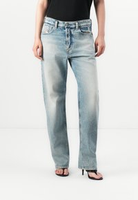 Persoon gekleed in lichtblauwe vervaagde recht-gesneden jeans en zwarte open teen hakken tegen een witte achtergrond.