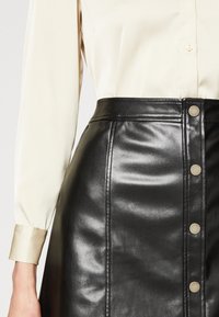 Juoda odinė sijonas su priekine sagų kišene, turintis lygią tekstūrą ir struktūruotą dizainą. Derinamas su šviesiu grietinėlės spalvos bluzonu.