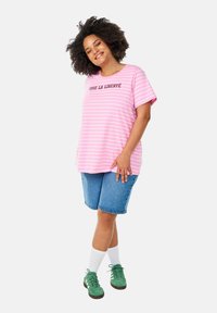 Roze en witte gestreepte T-shirt met de tekst "VIVE LA LIBERTÉ", gecombineerd met blauwe spijkerbroekshorts, witte sokken en groene sneakers.