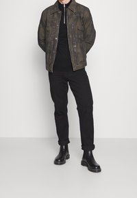 Giacca di jeans verde oliva con colletto zip, shirt nera a maniche lunghe, pantaloni neri con risvolto, e stivaletti neri alla caviglia con texture liscia.