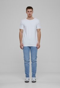 Fehér kerek nyakú póló, világoskék skinny farmernadrággal párosítva. A modellen fehér sportcipők vannak fekete és kék részletekkel. Egyszerű háttér.