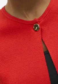 Punane õmmeldud kardigan on ümarate krae ja dekoratiivse kuldse nööbiga, millel on väike eesmine avamine, näidates musta sisekihti.