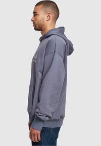 Sivé mikina s kapucňou a predným vreckom, s elastickými manžetami a logom na hrudi. Model zobrazený z boku, oblečený v džínsach.