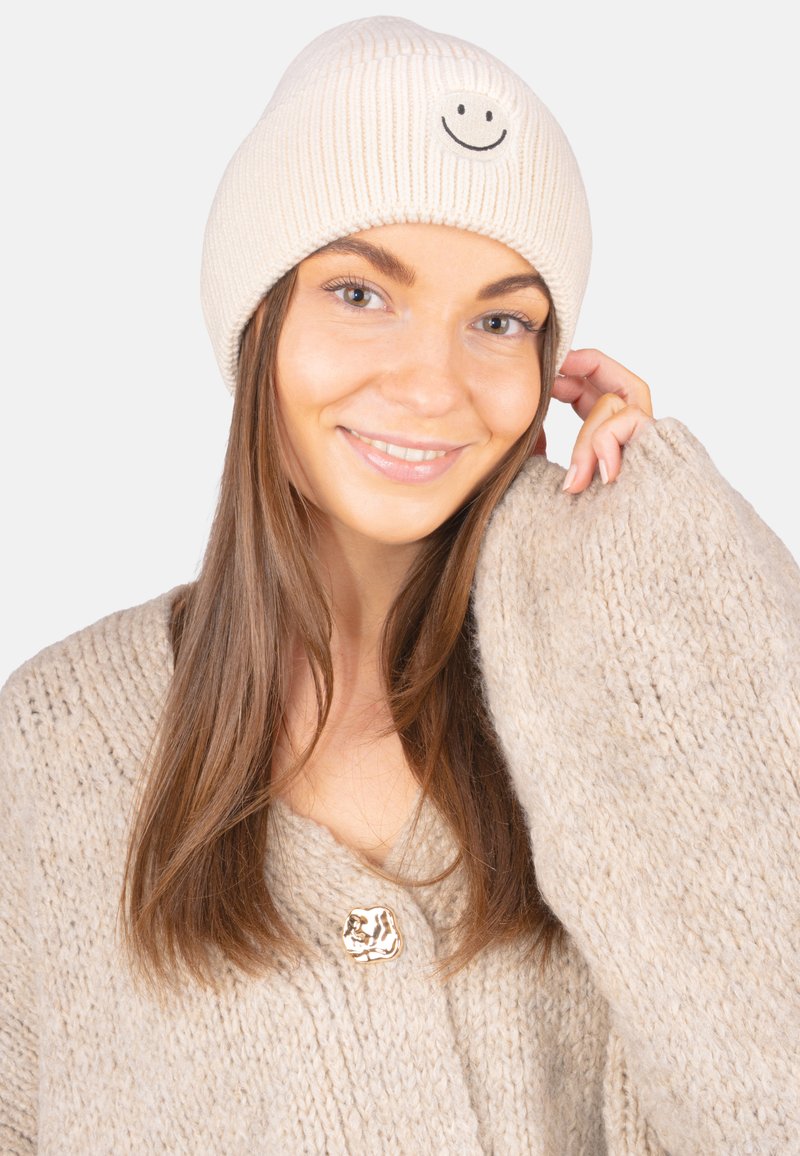 Bonnet en tricot côtelé crème avec un motif de smiley noir, porté avec un pull oversize texturé beige avec un bouton décoratif.