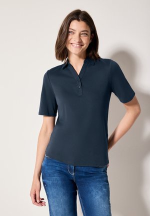 T-shirt polo bleu marine à manches courtes, avec un col boutonné et une texture lisse. Porté avec un jean bleu, présentant une coupe décontractée et des lignes épurées.
