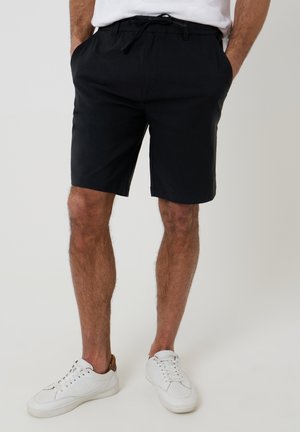 Mand iført sorte knælange shorts med hænderne i lommerne og hvide sneakers mod en ensfarvet baggrund.