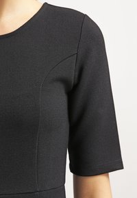 Černé šaty s krátkým rukávem, vyrobené z texturované tkaniny. Mají kulatý výstřih a zakřivené švy pro strukturovaný střih.