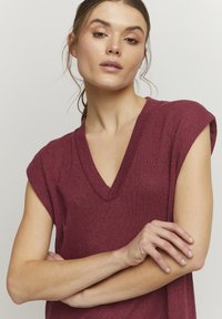 Haut en tricot bordeaux à col en V avec des manches courtes. Présente un tissu texturé et une coupe décontractée, mettant en avant un design léger.