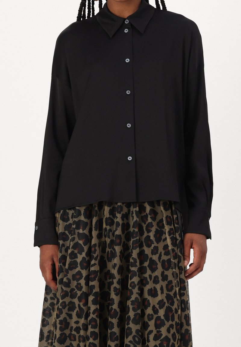 Chemise noire à manches longues avec col, en tissu lisse. Portée sur une jupe plissée à imprimé léopard vert et noir.