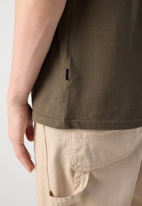 Oliven grønn T-skjorte laget av bomull, med en sideetikett. Kombinert med beige bukser som har en lappelomme og synlige sømmer.
