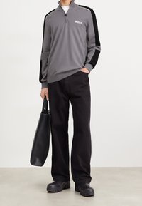 Mann trägt einen grauen BOSS-Pullover mit Viertelreißverschluss und schwarzen Streifen, schwarze Hose, klobige schwarze Schuhe und hält eine schwarze Ledertasche.