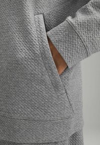 Szary teksturowany materiał ma kieszeń i ściągacz. Design jest casualowy, z diamentowym wzorem dodającym wizualnego interesu.