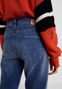 Jean en denim bleu avec une taille haute, présentant un patch en cuir marron, des détails usés et deux poches arrière. Pull en tricot rouge avec des accents noirs.