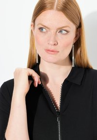 Czarna bluza na zamek z dekoracyjnymi koralikami wzdłuż zamka, w komplecie z wiszącymi srebrnymi kolczykami. Gładki materiał i dopasowany krój.